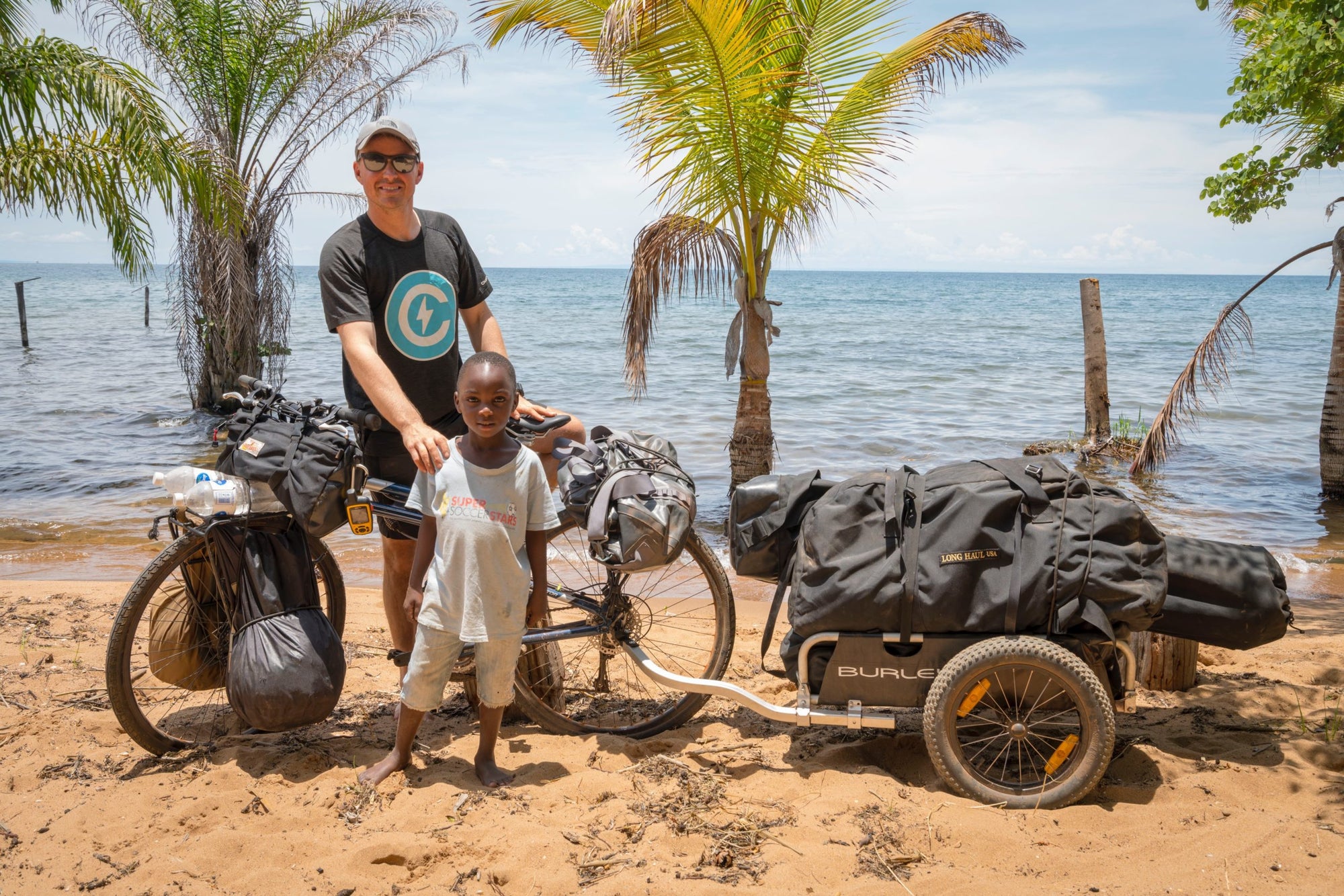 man standing with child in frontof bike and fully loaded cargo trailer on a sandy shore. lake and palm trees in background