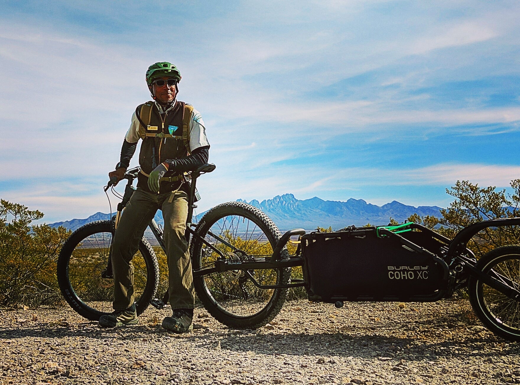 man leaning against bicycle with cargo bike trailer attached on a dirt path. blue sky and clouds in background