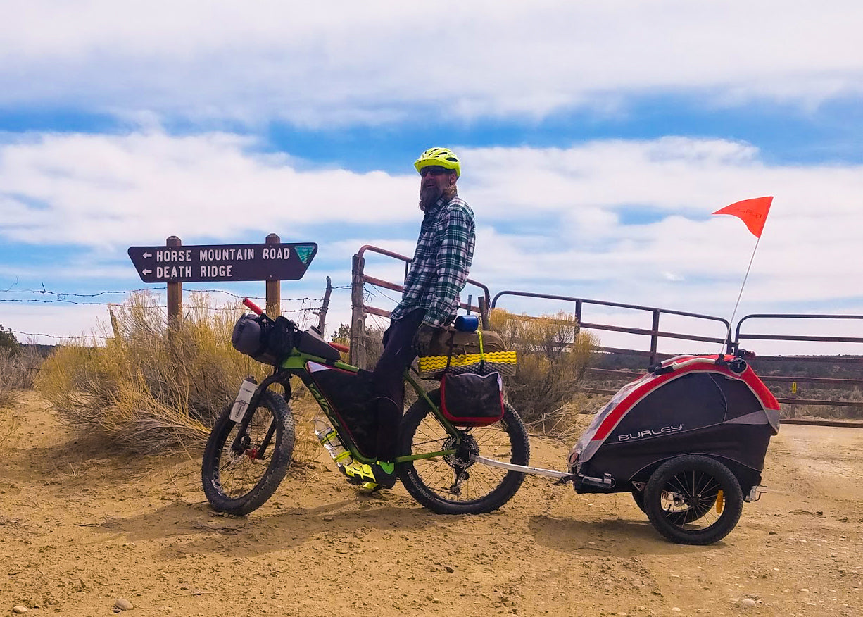 man sittingon bike with kids bike trailer attached on dirt road with blue sky in background