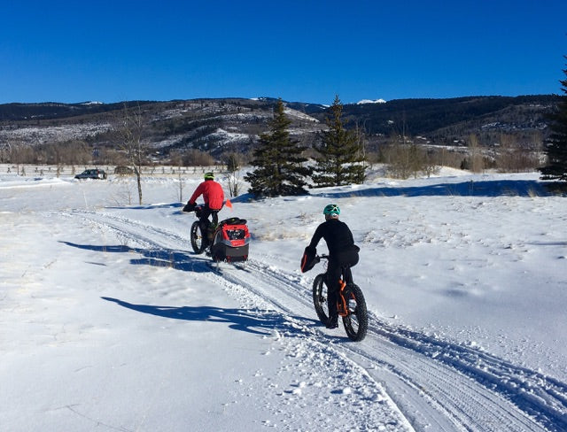 two people biking on snowy covered road. one towing kids bike trailer