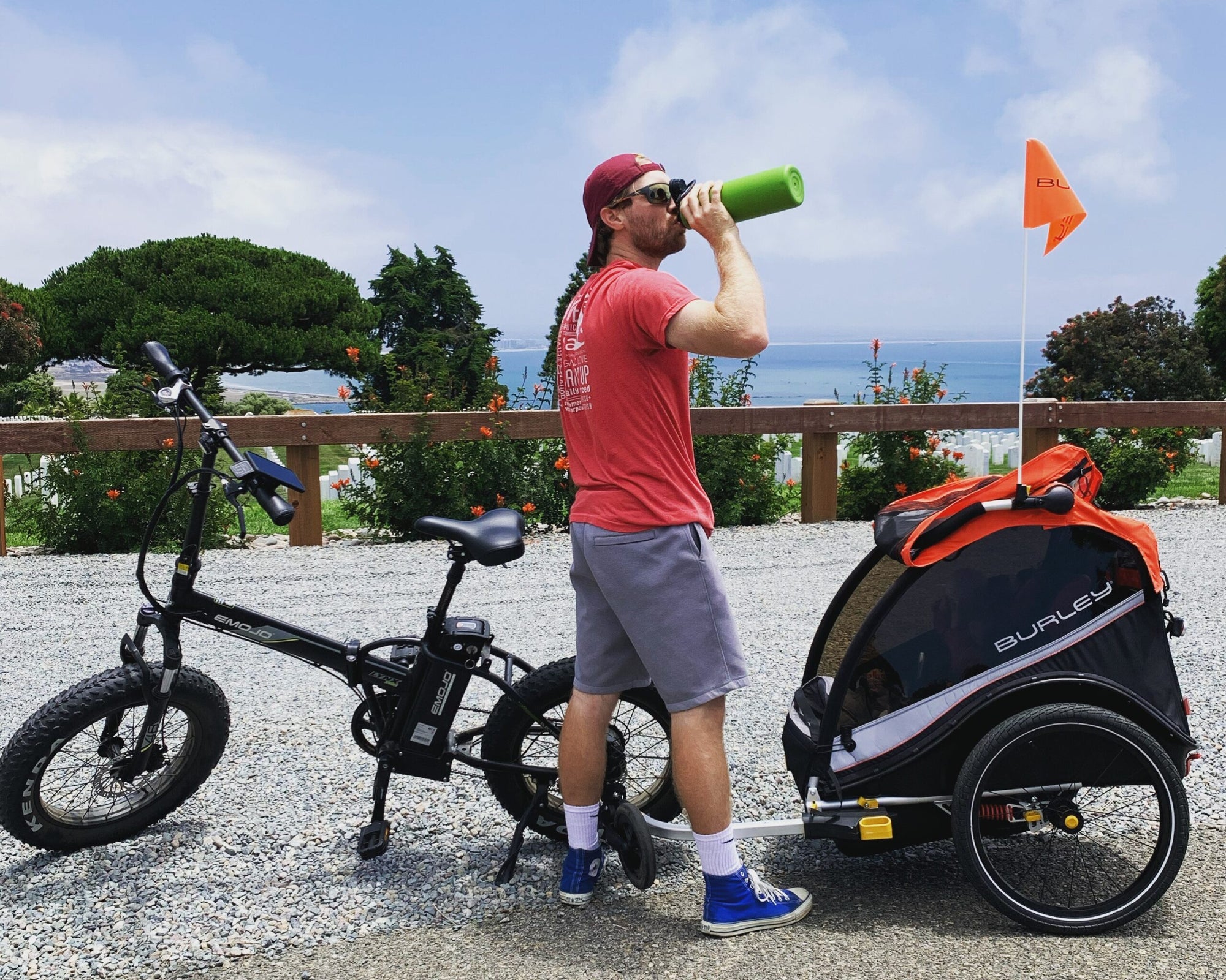 man drinks from water bottle in front of bike with kids bike trailer on teh back. ocean in background