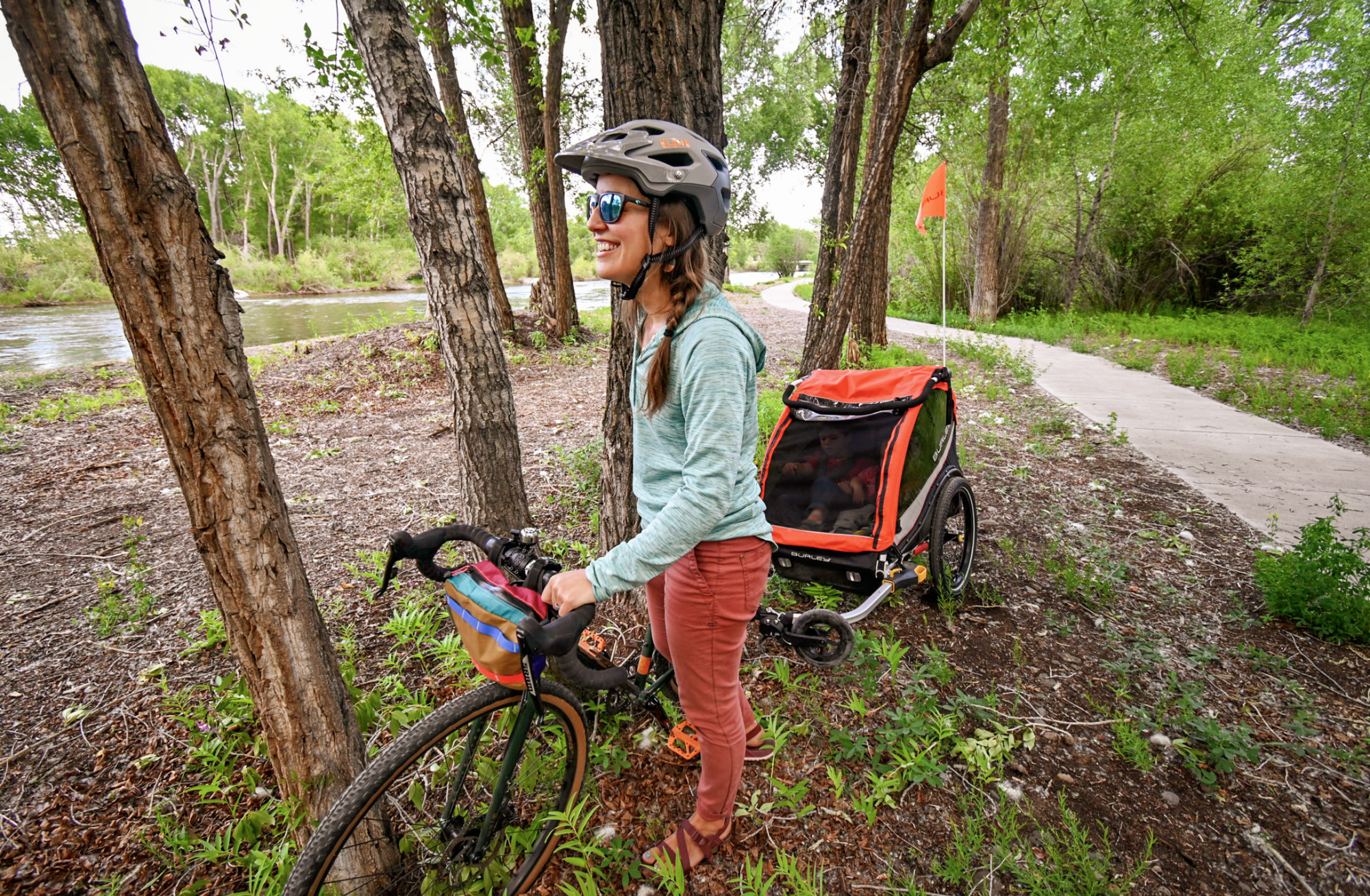 woman standing among trees and holding onto bike with kid bike trailer attached