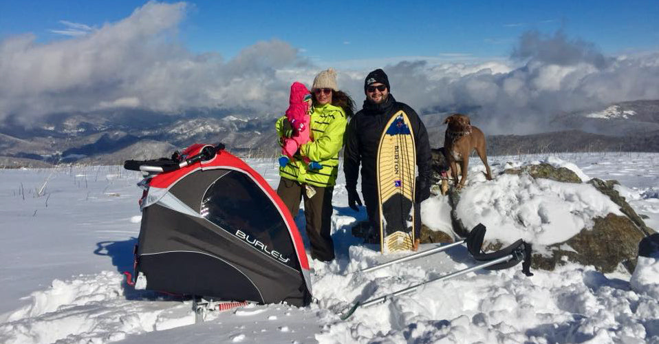man and woman standing in snow with snowboards and kids bike trailer. woman holding toddler