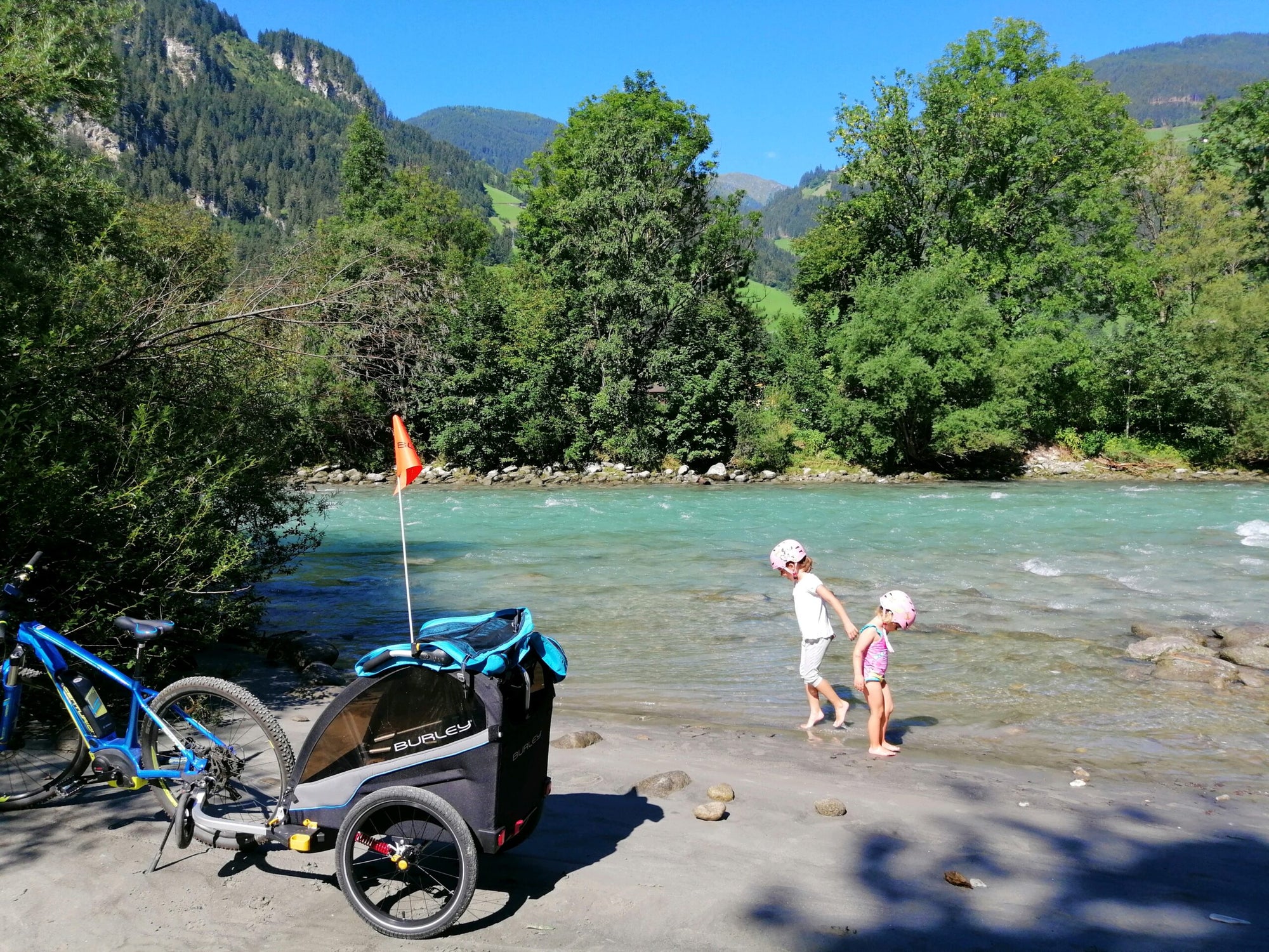two young girls wading in river. bicycle with kid bike trailer attached parked alongside river