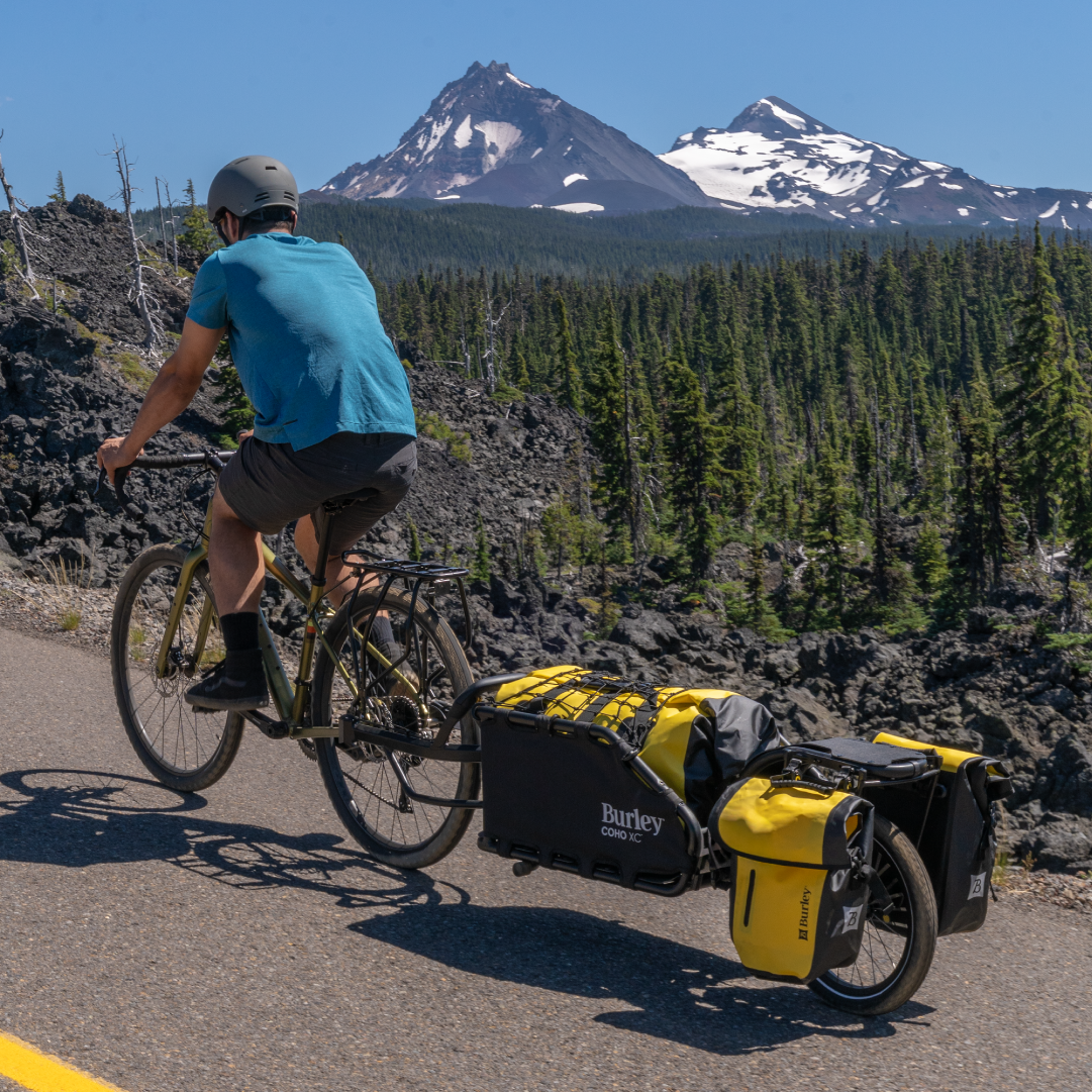 Person cycling with a Coho XC Cargo Trailer on a road with mountains in the background