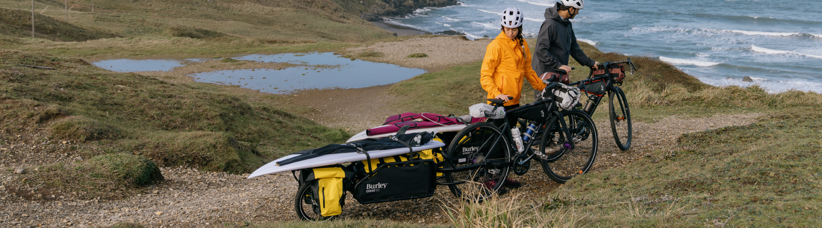 Two people with Coho XC Cargo Bike Trailer hauling surfboards on a grassy hill near a coastal area.