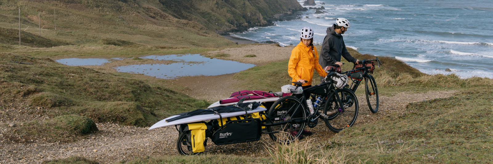 Two people with Coho XC Cargo Bike Trailer hauling surfboards on a grassy hill near a coastal area.