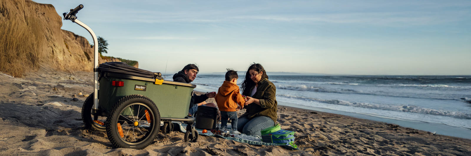 Family sitting by a Hopper Bike Trailer & Cargo Wagon on a beach with ocean waves in the background