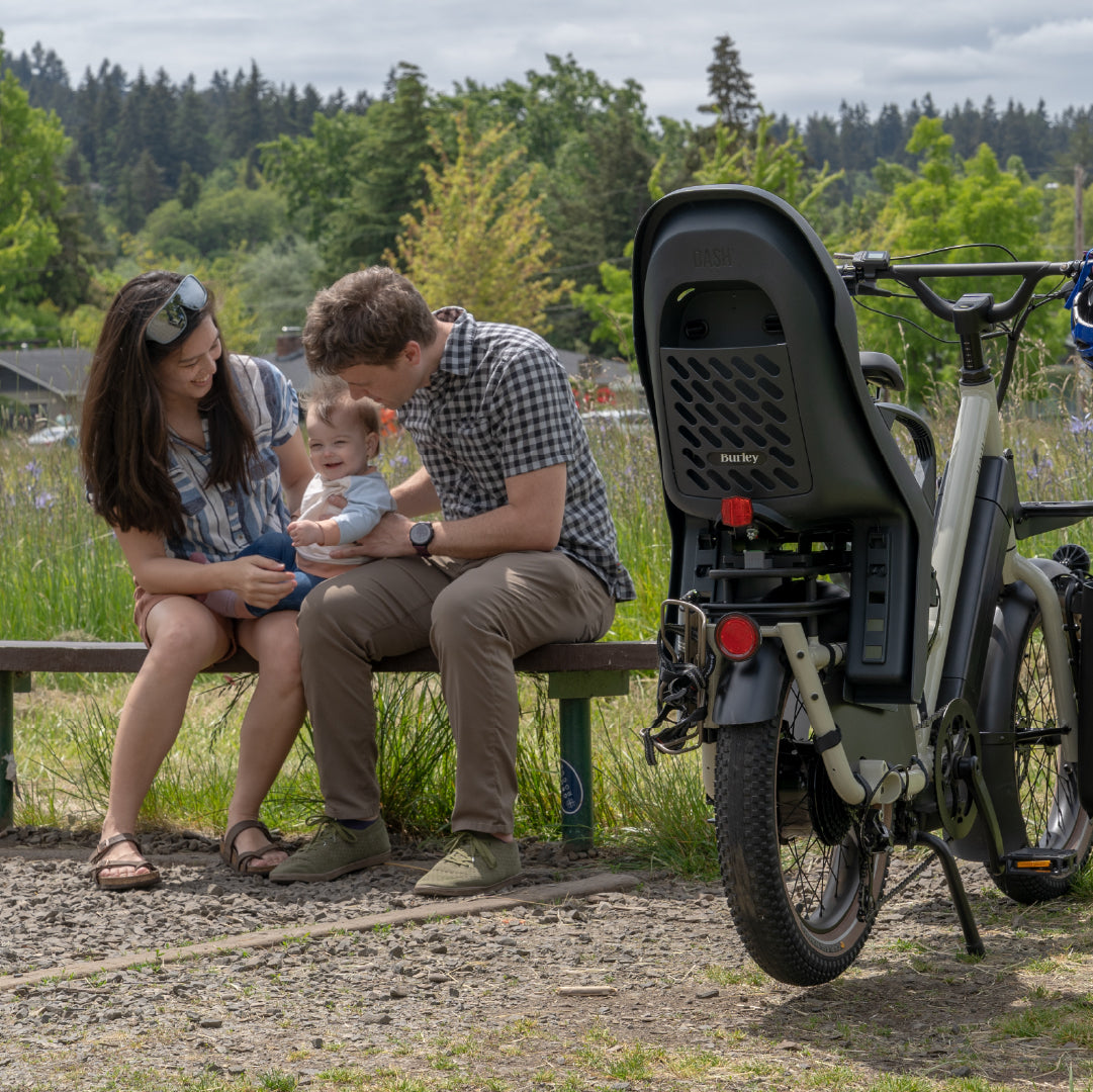 Family with a child sitting on a bench next to a cargo bike with a Burley Dash child seat in a natural setting.