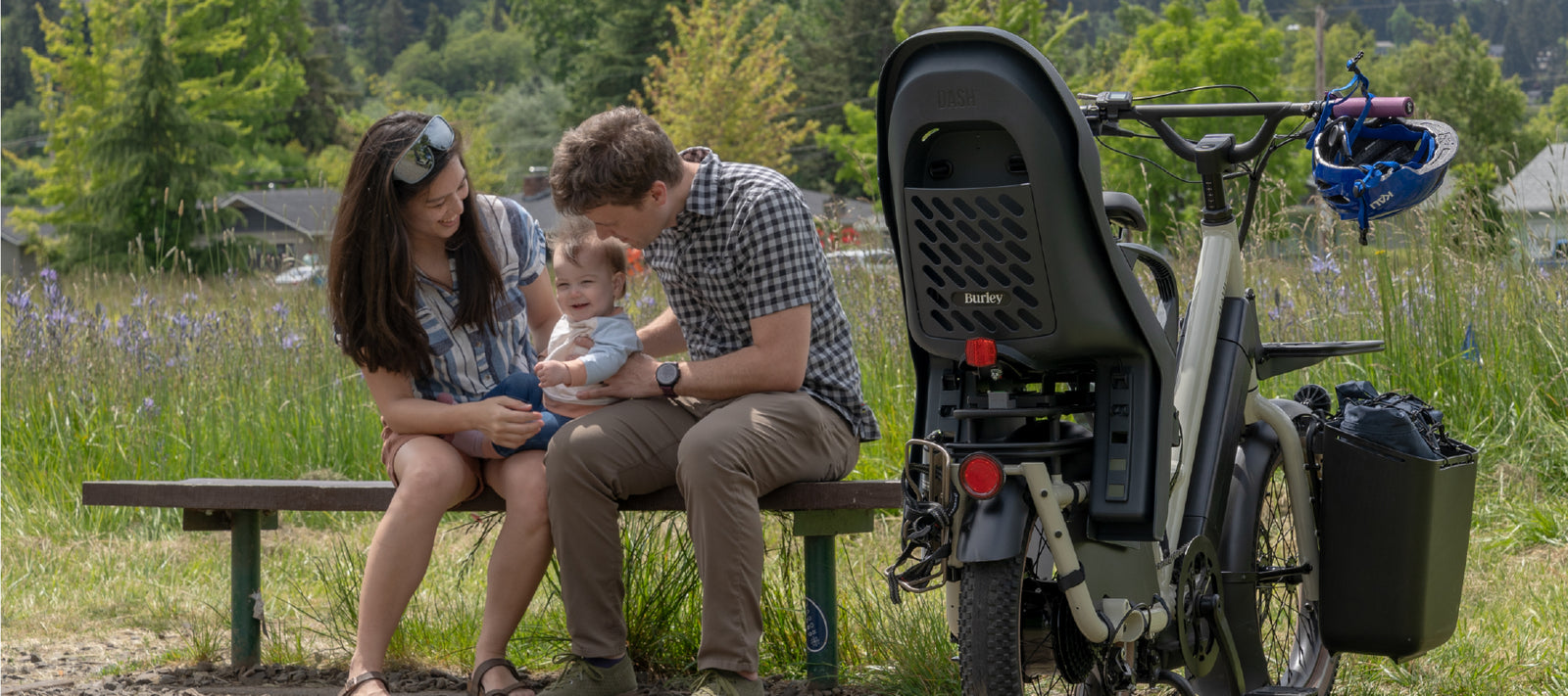Family with a child sitting on a bench next to a cargo bike with a Burley Dash child seat in a natural setting.