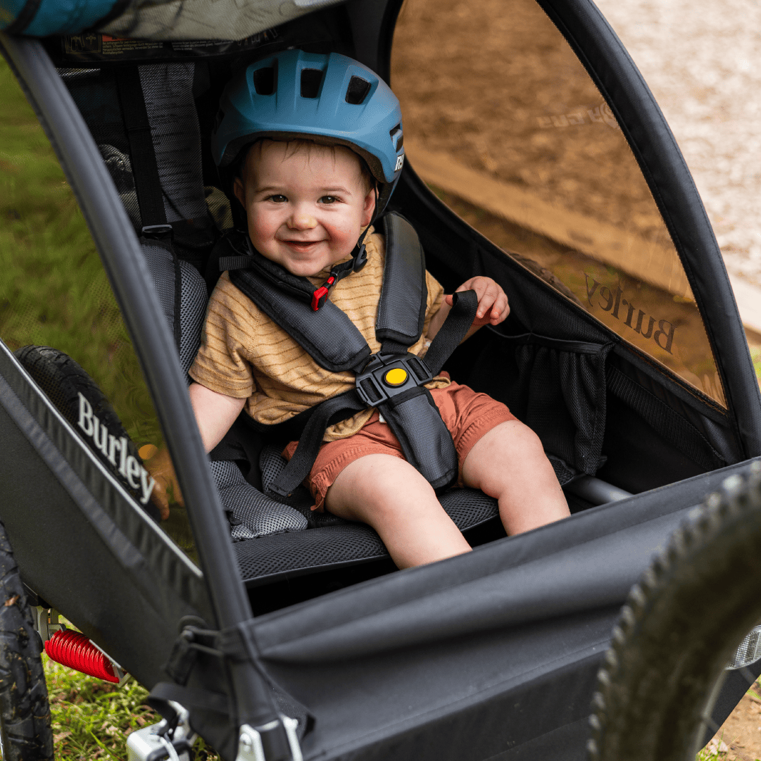 Child smiling in a Burley Encore X bike trailer with a helmet, sitting outdoors.